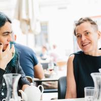 Susanne Kirchmayr aka Electric Indigo und Angélica Castelló im Interview. Sie sitzen an einem Tisch, Susanne Kirchmayr hat einen Sidecut, trägt ein schwarzes Oberteil und lacht. Susanne Kirchmayr hat schwarz-grüne Haare. Foto: Niko Havranek