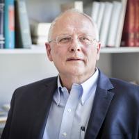 Ein Foto von Stefan Hopman. Er trägt ein hellblaues Hemd und einen dunkelblauen Anzug. Er hat eine Halbglatze und trägt eine dünne Brille. Er sieht konzentriert aus. 