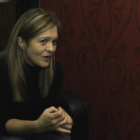 Foto von Barbara Eder in einem schwarzen Sessel vor einer roten, gemusterten Wand. Barbara Eder schaut konzentriert, Interviewsituation.